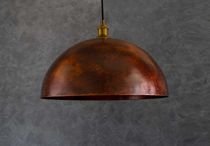 An oxidized smooth copper dome pendant light with a textured metallic finish, suspended by a black cord and set against a dark gray textured wall.