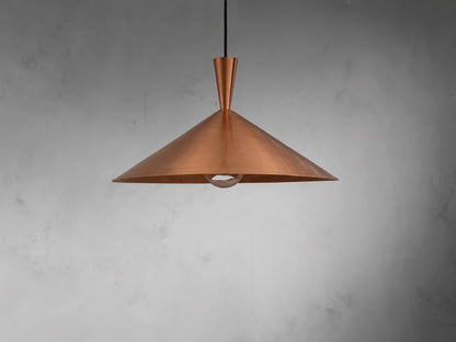A copper pendant light with a wide, shallow conical shade made of brushed copper, suspended from a black cord against a textured gray wall.