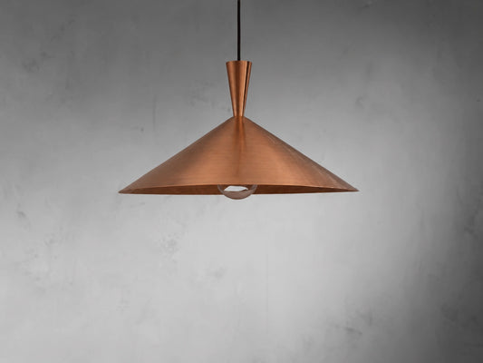 A copper pendant light with a wide, shallow conical shade made of brushed copper, suspended from a black cord against a textured gray wall.